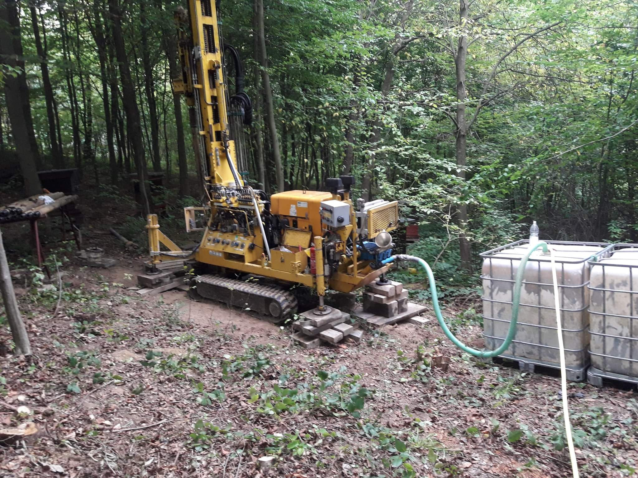 Żółta wiertnica na gąsienicach, ustawiona na betonowych płytach w zalesionym terenie, z podłączonymi zbiornikami na wodę, przygotowana do wiercenia.