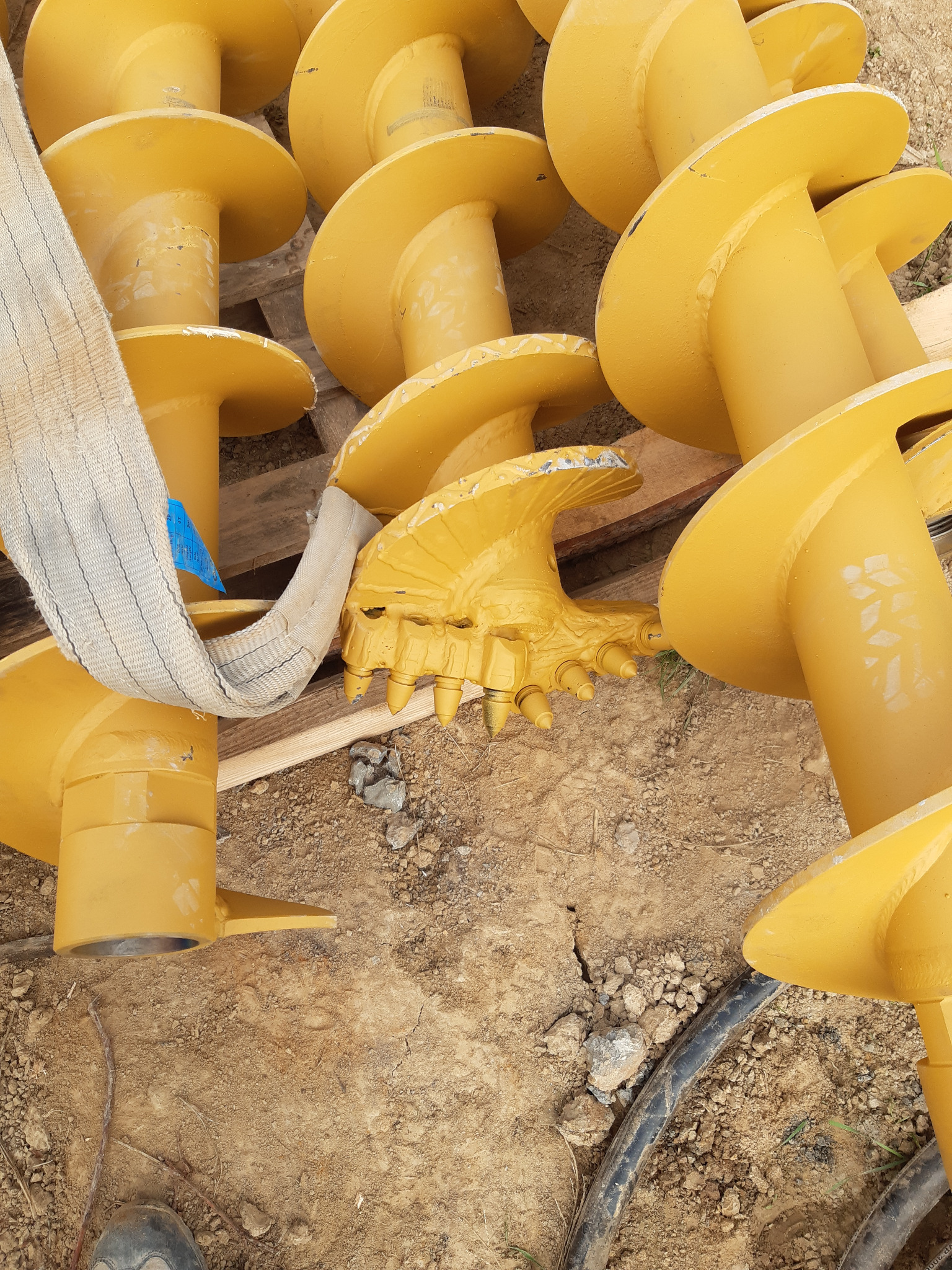 Żółte świdry do wiertnicy ułożone na drewnianej palecie i ziemi, jeden ze świdrów ma dodatkowe zęby do wiercenia w twardym gruncie, obok leży pas transportowy.