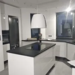 Nowoczesna biała kuchnia z wyspą i czarnym blatem, oświetlona wiszącą lampą z okapem, widoczny sprzęt AGD.