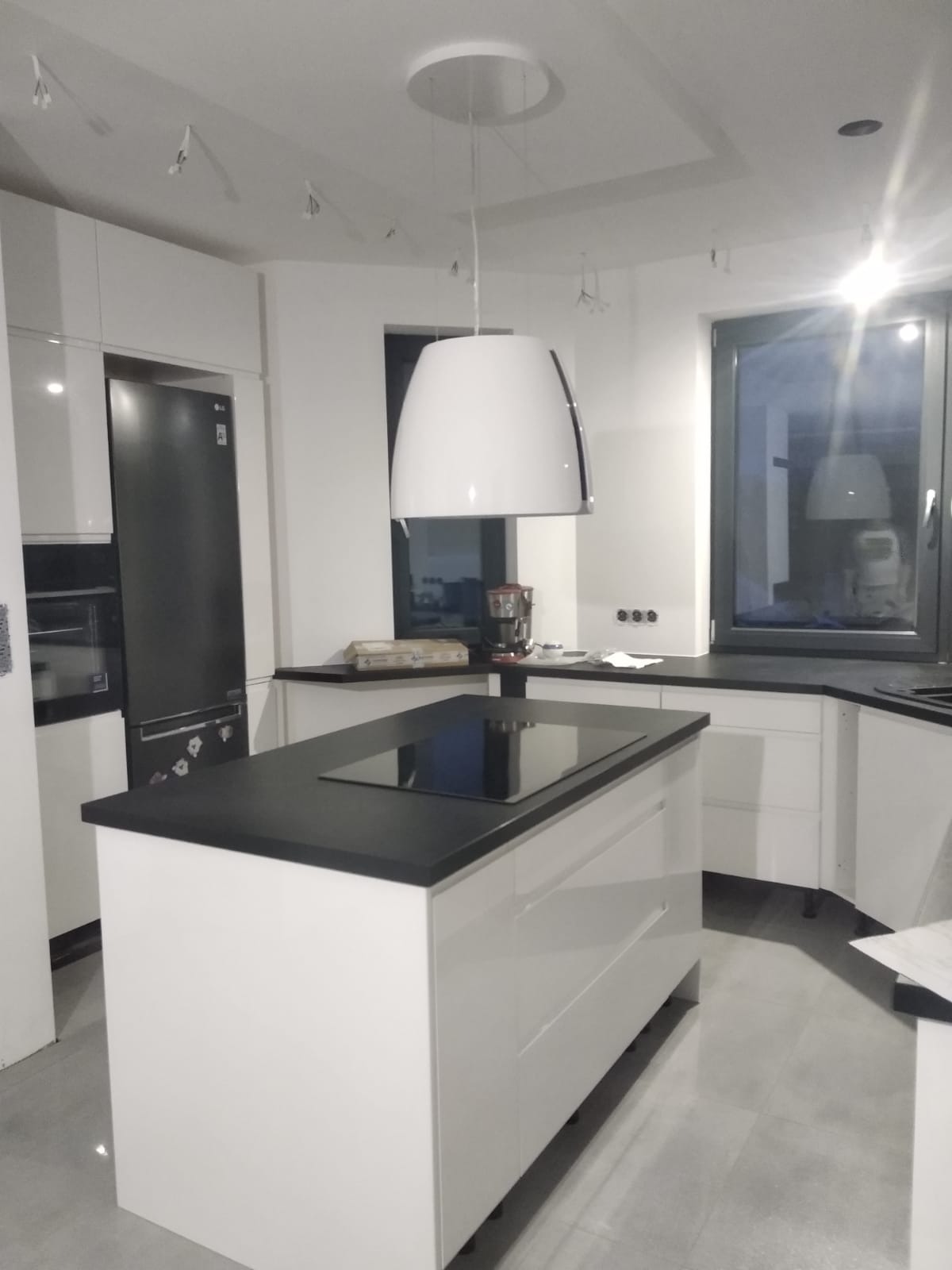 Nowoczesna biała kuchnia z wyspą i czarnym blatem, oświetlona wiszącą lampą z okapem, widoczny sprzęt AGD.