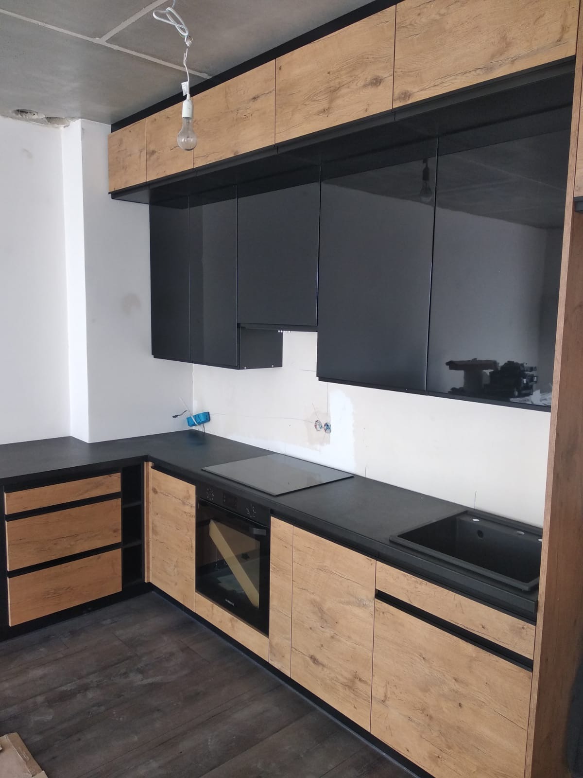 Częściowo zamontowana kuchnia z czarnymi, błyszczącymi szafkami wiszącymi i dolnymi szafkami z drewnopodobnym wykończeniem, czarnym blatem i niedokończonym sufitem.
