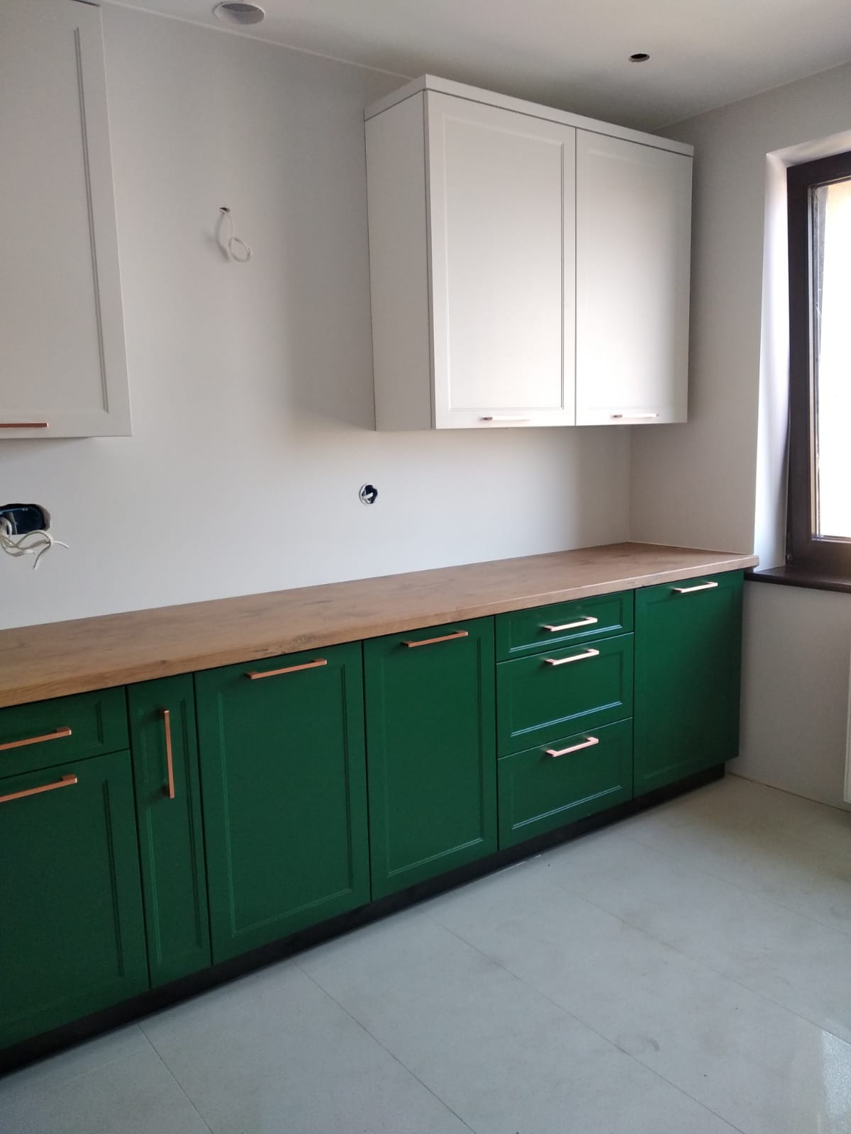 Zielone szafki kuchenne z drewnianym blatem i miedzianymi uchwytami w Pszczynie, białe szafki wiszące, widoczne przygotowania do montażu oświetlenia.