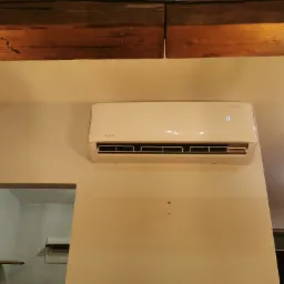 Biały klimatyzator Rotenso na ścianie w pomieszczeniu z drewnianymi belkami stropowymi i dekoracyjną lampą sznurową.