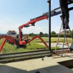 Montaż okna z użyciem mini żurawia pająka na zewnątrz budynku, widok z wnętrza przez ramę okienną, pracownik na drabinie w tle.