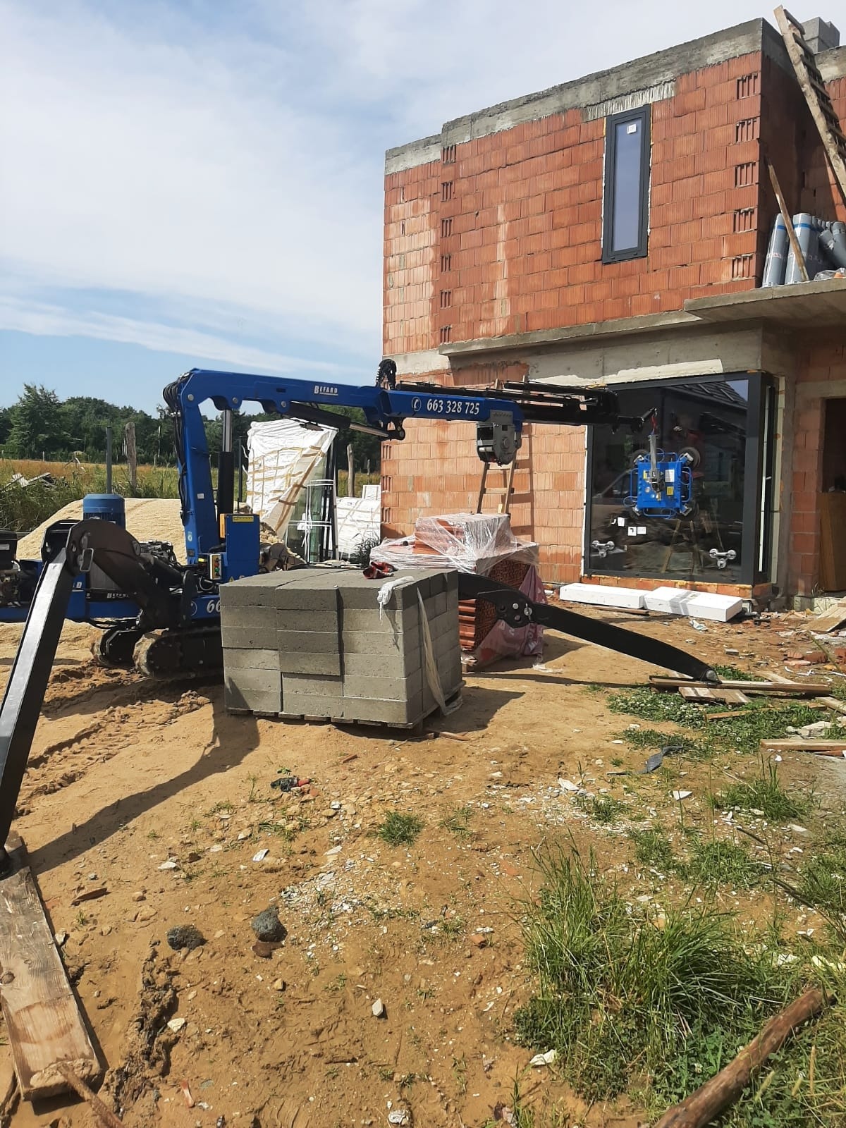 Niebieski mini-dźwig gąsienicowy podnosi szybę okienną z przyssawkami podczas montażu w budynku z czerwonej cegły. Na pierwszym planie ułożone bloczki betonowe.