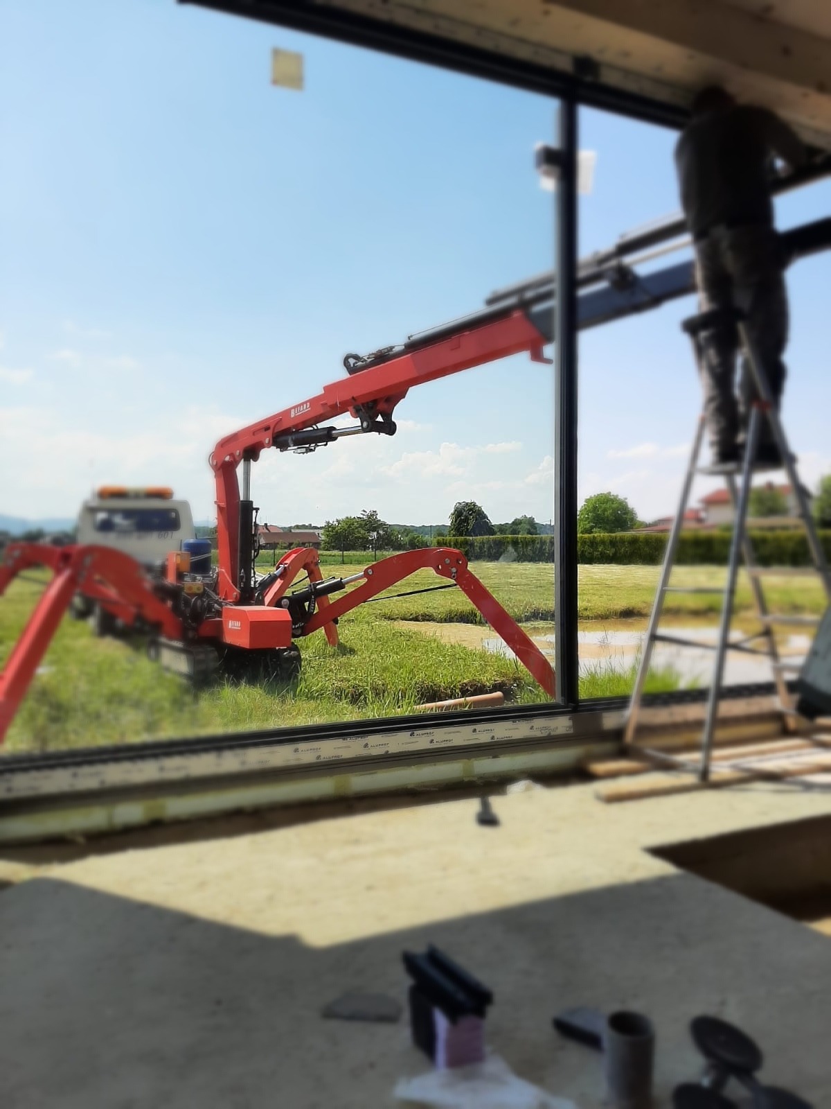 Montaż okna z użyciem mini żurawia pająka na zewnątrz budynku, widok z wnętrza przez ramę okienną, pracownik na drabinie w tle.