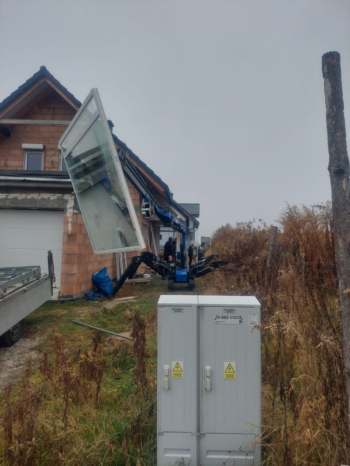 Montaż okna w Jankowicach za pomocą małego dźwigu na gąsienicach, widoczna surowa elewacja budynku w trakcie budowy.