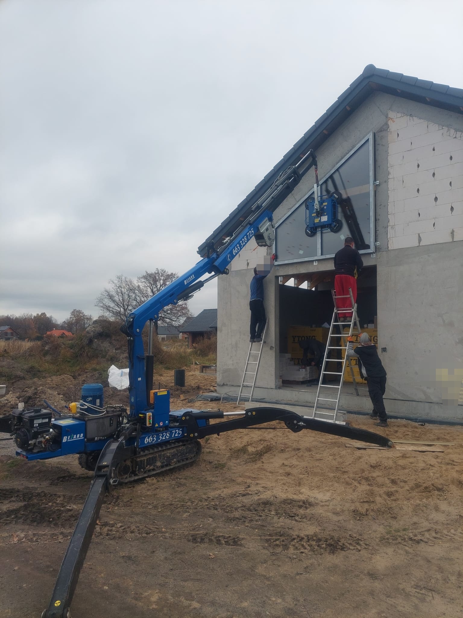 Montaż okna w surowym budynku z użyciem mini-dźwigu gąsienicowego i przyssawek próżniowych, pracownicy na drabinach, widoczne bloczki Ytong.