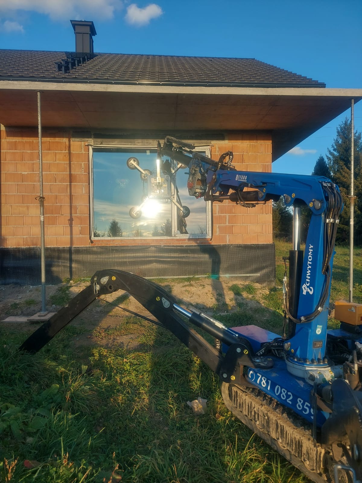 Niebieski, gąsienicowy mini dźwig z przyssawkami montuje okno w surowym budynku z czerwonej cegły, w tle widoczne drzewa i błękitne niebo.