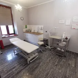 Jasne wnętrze gabinetu lekarskiego z kozetką, szafkami i certyfikatami na ścianie. Widoczne wyposażenie medyczne i lampa. Podłoga w odcieniach szarości.