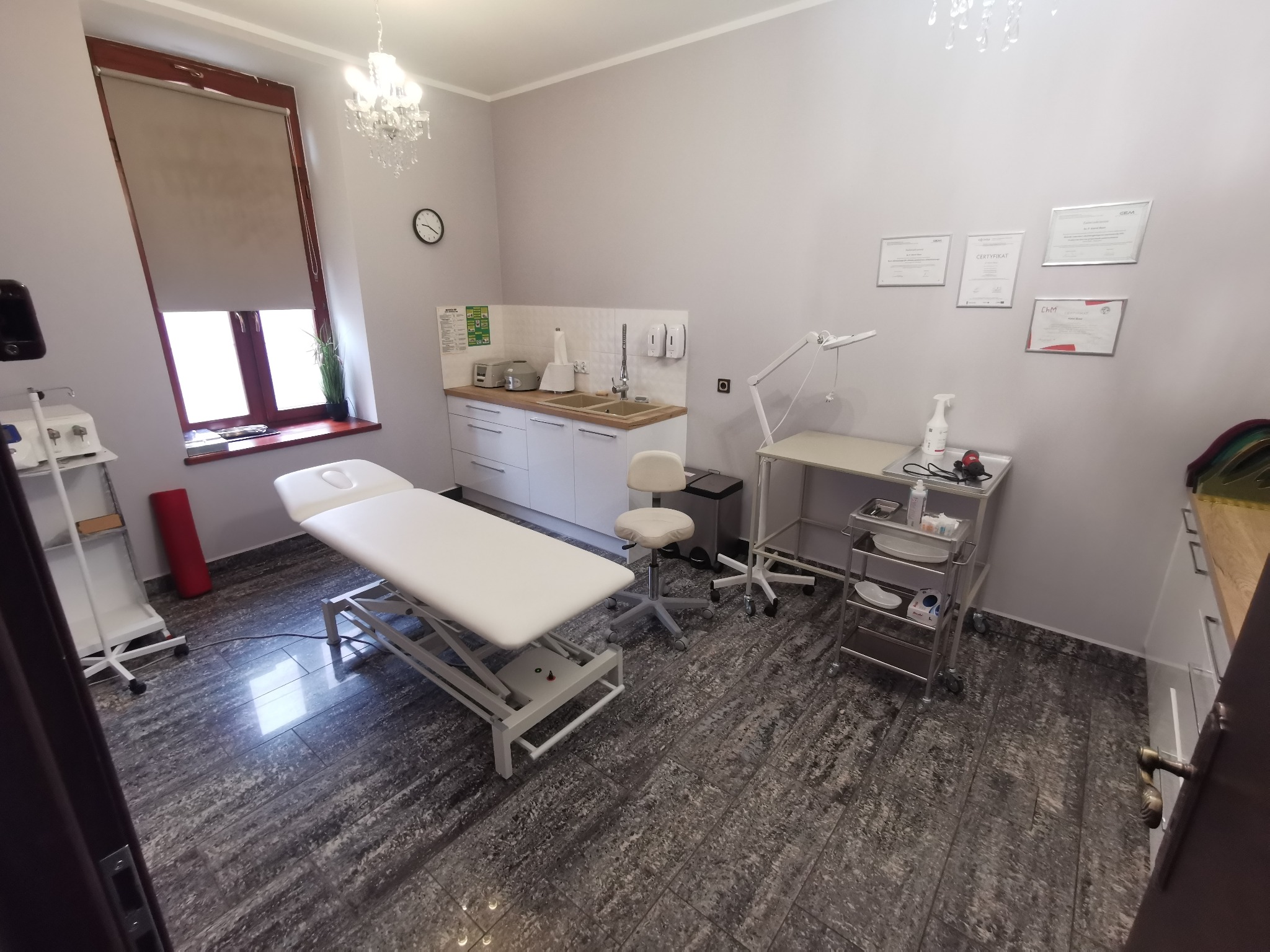 Jasne wnętrze gabinetu lekarskiego z kozetką, szafkami i certyfikatami na ścianie. Widoczne wyposażenie medyczne i lampa. Podłoga w odcieniach szarości.