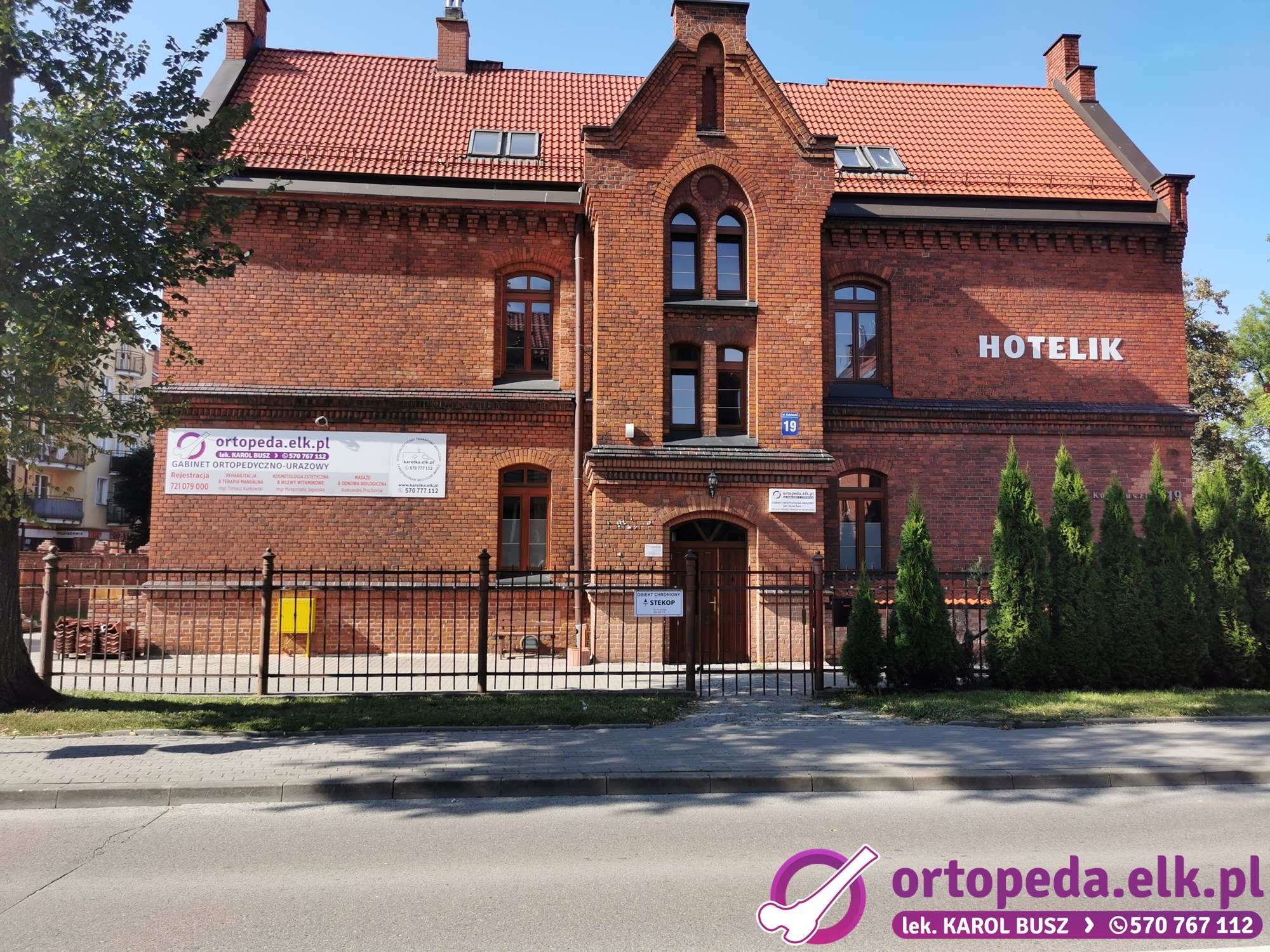 Fasada budynku ortopedii z szyldem 'ortopeda.elk.pl', numerem 19 i napisem 'HOTELIK' na tle błękitnego nieba. Widoczny chodnik i fragment ulicy.