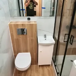 Nowoczesna łazienka z białymi płytkami imitującymi marmur, drewnopodobną zabudową toalety, kabiną prysznicową z czarnymi ramami i odbiciem fotografa w lustrze.