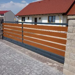 Przesuwna brama wjazdowa z drewnopodobnymi panelami, osadzona między kamiennymi słupkami na tle domu z czerwoną dachówką i brukowanego podjazdu.