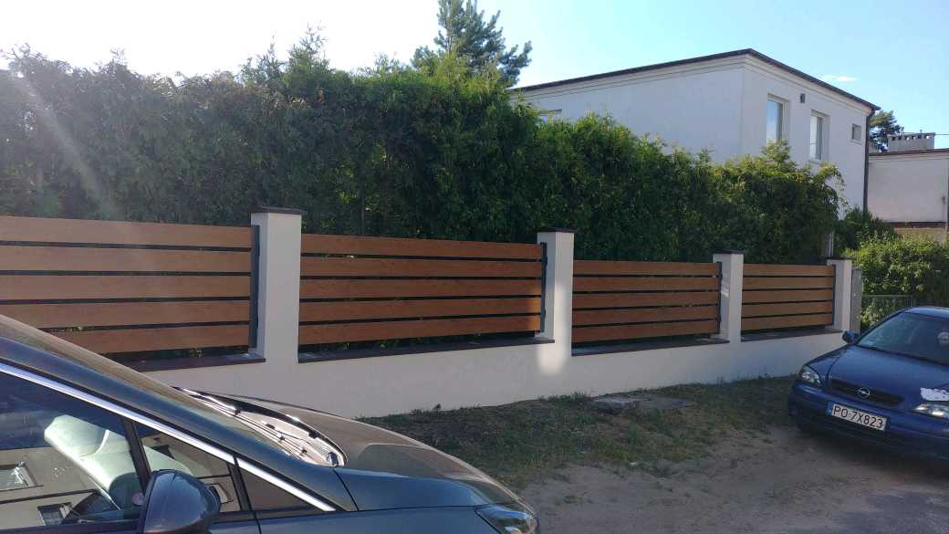 Nowoczesne ogrodzenie drewnopodobne z poziomych paneli w kolorze jasnego drewna, osadzone na białym murku z filarami, z widocznymi samochodami na pierwszym planie i zielenią w tle.