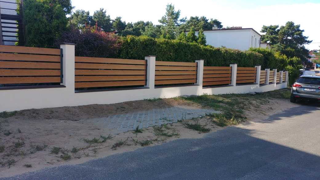 Nowoczesne ogrodzenie drewnopodobne z poziomych paneli na niskiej, białej podmurówce i białych słupkach, widok z zewnątrz, z fragmentem ulicy i samochodem w tle.