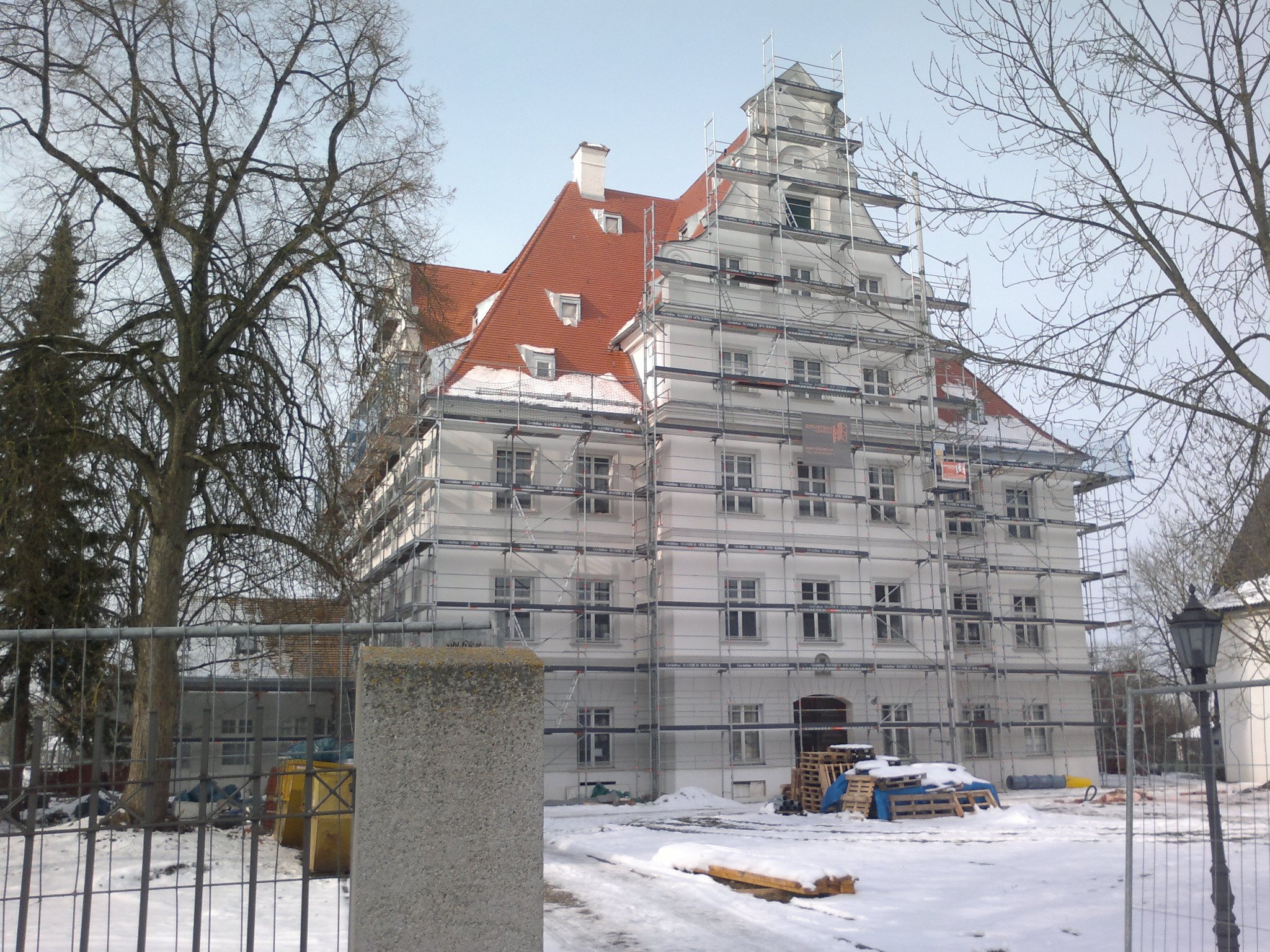 Duży, biały budynek w rusztowaniach z pomarańczową dachówką, częściowo pokrytą śniegiem, widoczny zimą, z drzewami bez liści w tle.