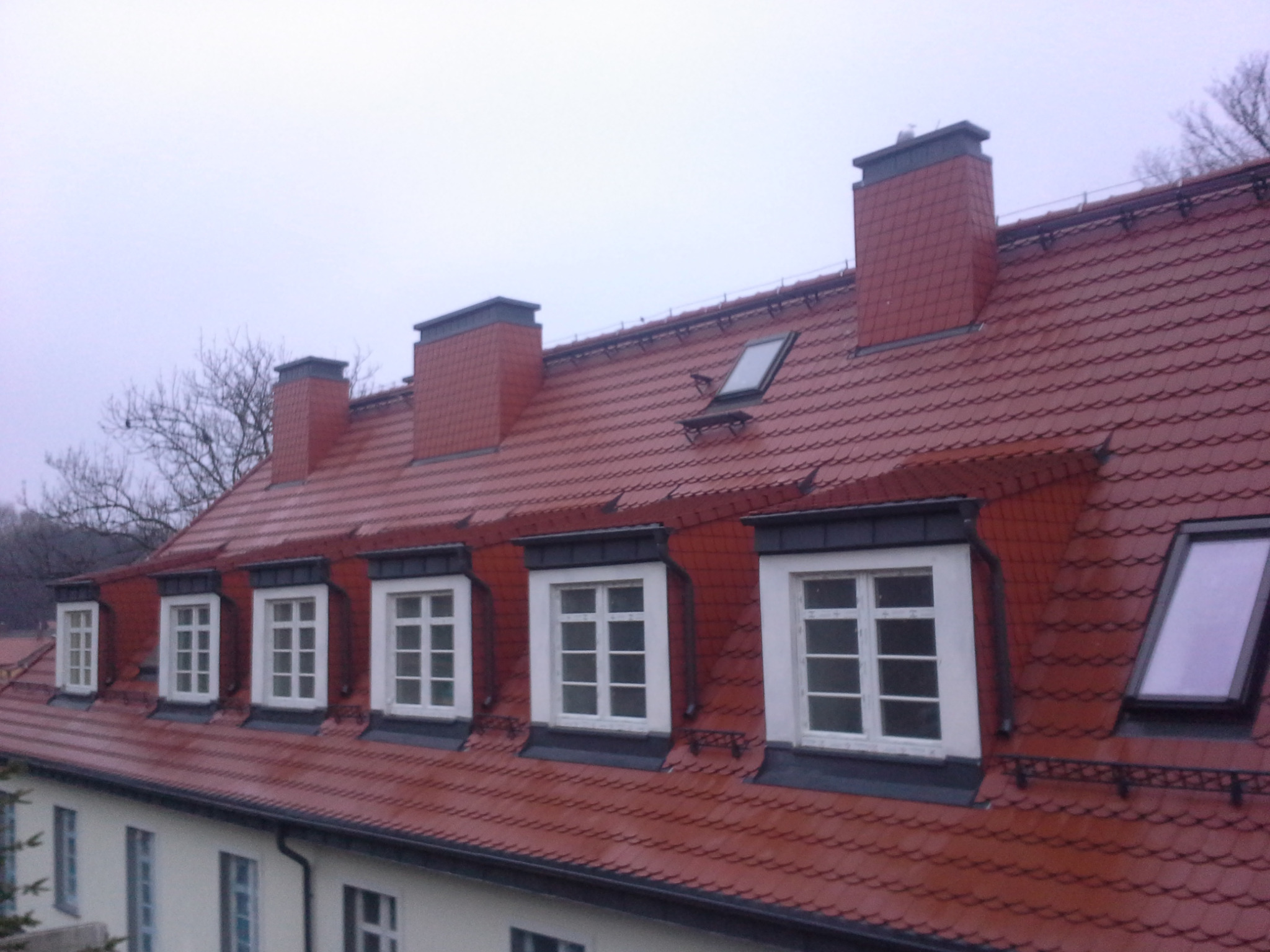 Czerwony dach z dachówką karpiówką, trzema kominami, oknami dachowymi i lukarnami, widziany z dołu w pochmurny dzień.