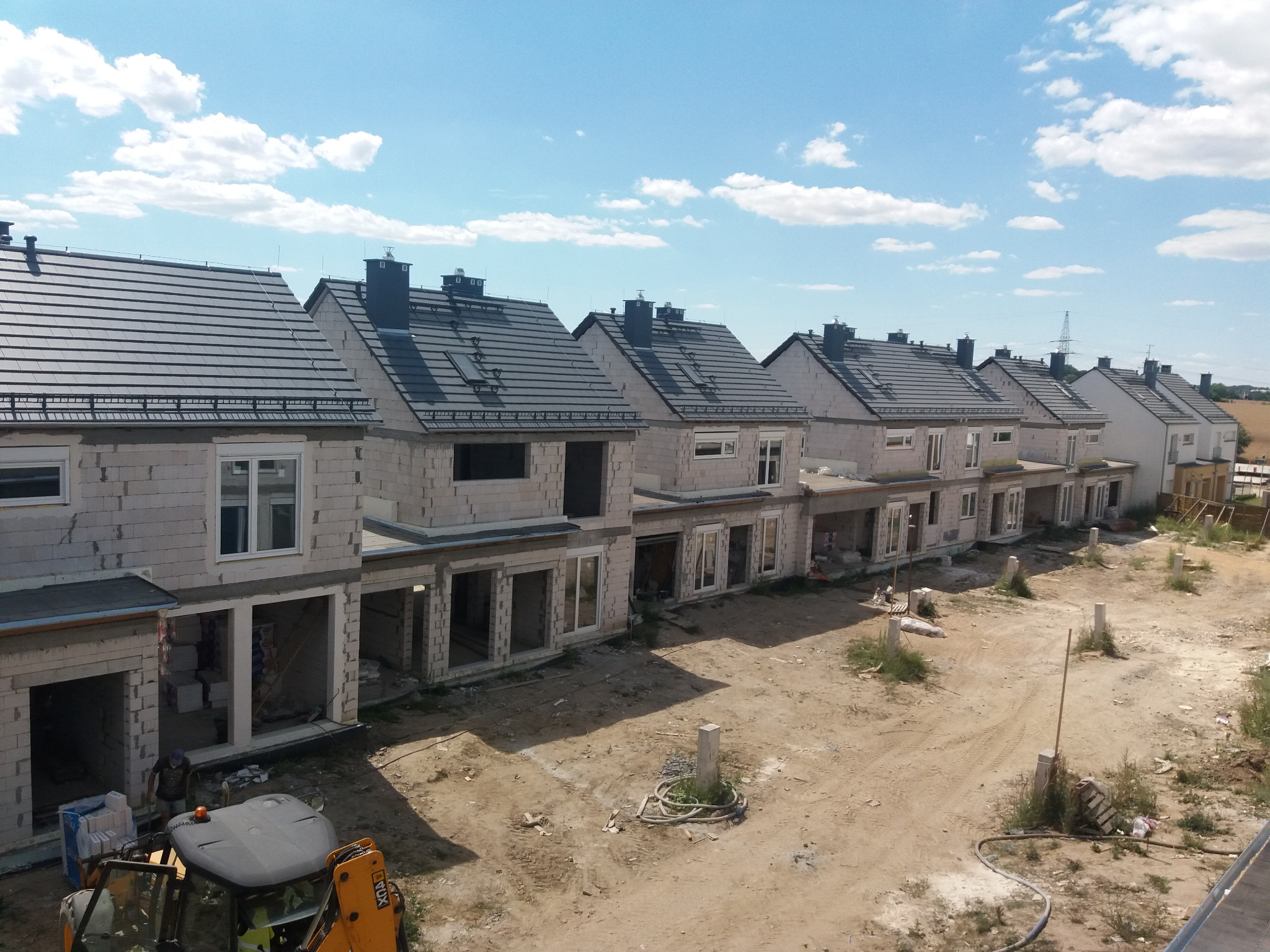 Rząd domów w trakcie budowy z ciemnoszarą dachówką, widoczny z góry, z placem budowy i minikoparką na pierwszym planie.
