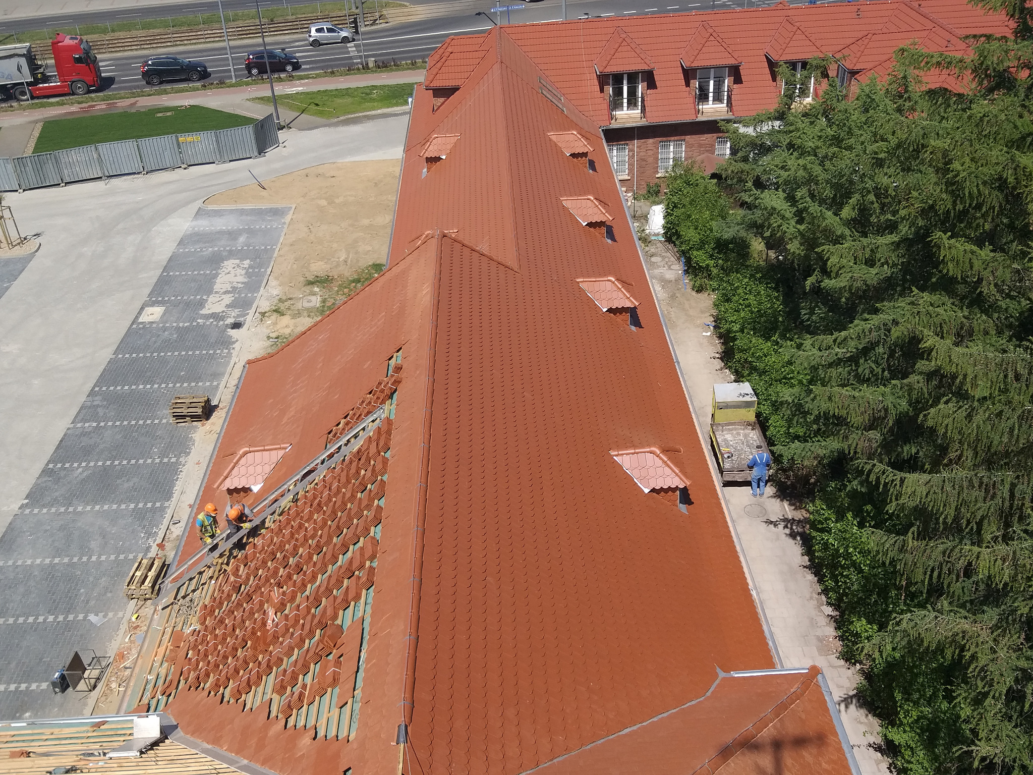 Widok z góry na częściowo pokryty czerwoną dachówką dach budynku, dwóch robotników w kaskach i kamizelkach odblaskowych układających dachówkę oraz fragment dachu sąsiedniego budynku, drzewa i chodnik.