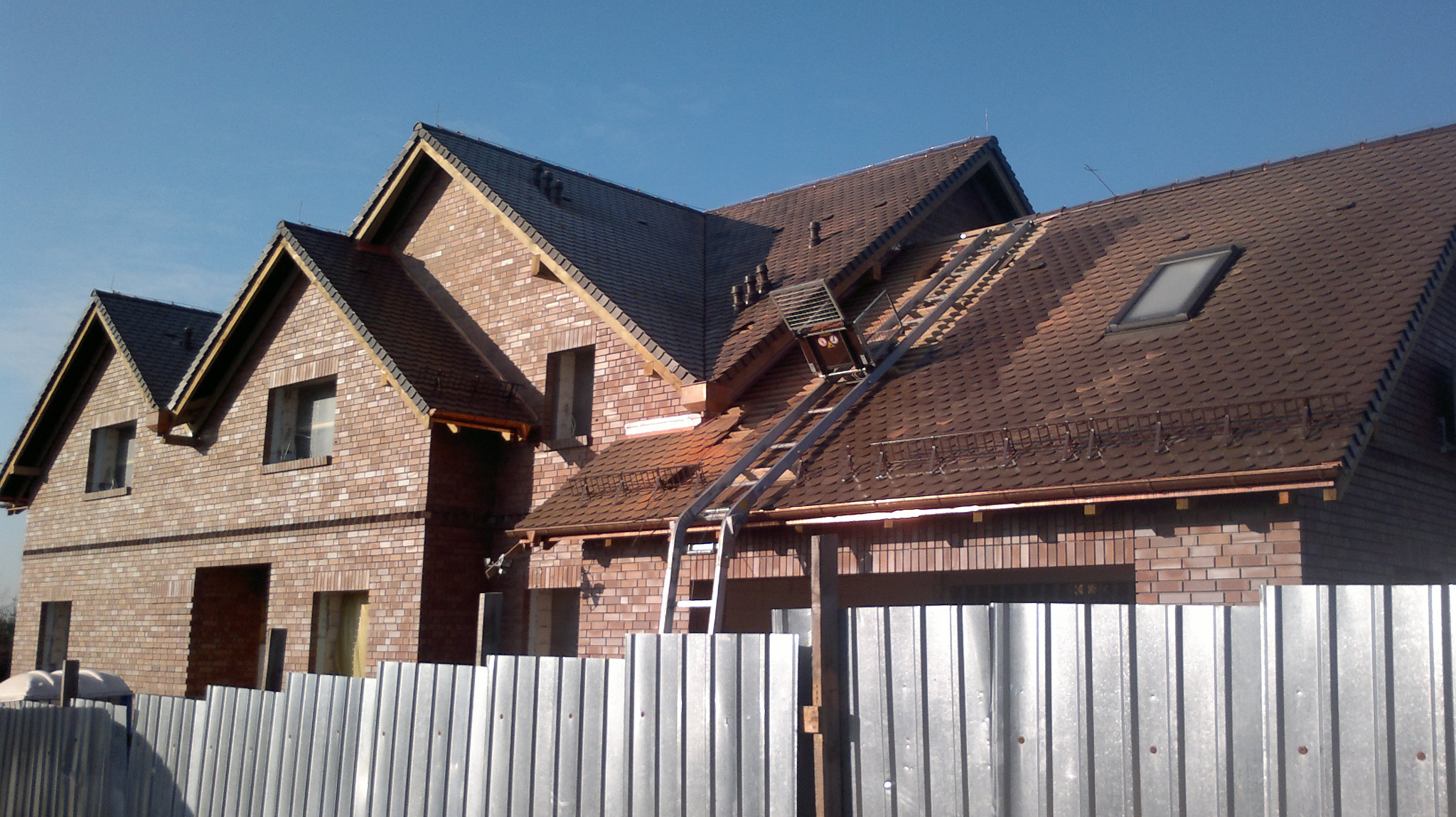 Dom w trakcie budowy z częściowo pokrytym dachem brązową dachówką, widoczna drabina dekarska i rynny miedziane, w tle błękitne niebo.