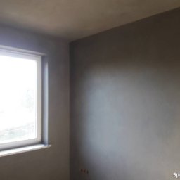 Usługi ogólnobudowlane Łukasz Grupa - Świeżo nałożony, szary tynk na ścianach w pokoju z oknem, widoczne niedoskonałości faktury przed dalszą obróbką.