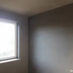 Świeżo nałożony, szary tynk na ścianach w pokoju z oknem, widoczne niedoskonałości faktury przed dalszą obróbką.