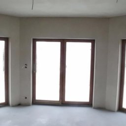 Usługi ogólnobudowlane Łukasz Grupa - Pokój z trzema oknami w drewnianych ramach, ściany po nałożeniu gładzi, widoczne wystające kable i puszki elektryczne.