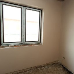 Usługi ogólnobudowlane Łukasz Grupa - Pokój w trakcie prac wykończeniowych, ściany świeżo pokryte gładzią, okno z brązową ramą, surowa podłoga przed położeniem posadzki, widoczne ślady po instalacjach elektrycznych.