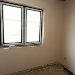 Pokój w trakcie prac wykończeniowych, ściany świeżo pokryte gładzią, okno z brązową ramą, surowa podłoga przed położeniem posadzki, widoczne ślady po instalacjach elektrycznych.