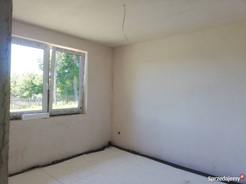 Surowe wnętrze pokoju z oknem, ściany po wstępnym przygotowaniu, podłoga zabezpieczona folią, widoczne niedoskonałości tynku i wystający kabel z sufitu.