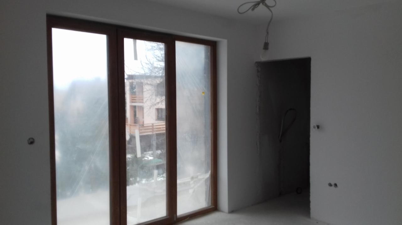 Surowe wnętrze po szpachlowaniu, z widocznymi przewodami elektrycznymi i dużym oknem z drewnianą ramą, przez które widać budynek w tle.