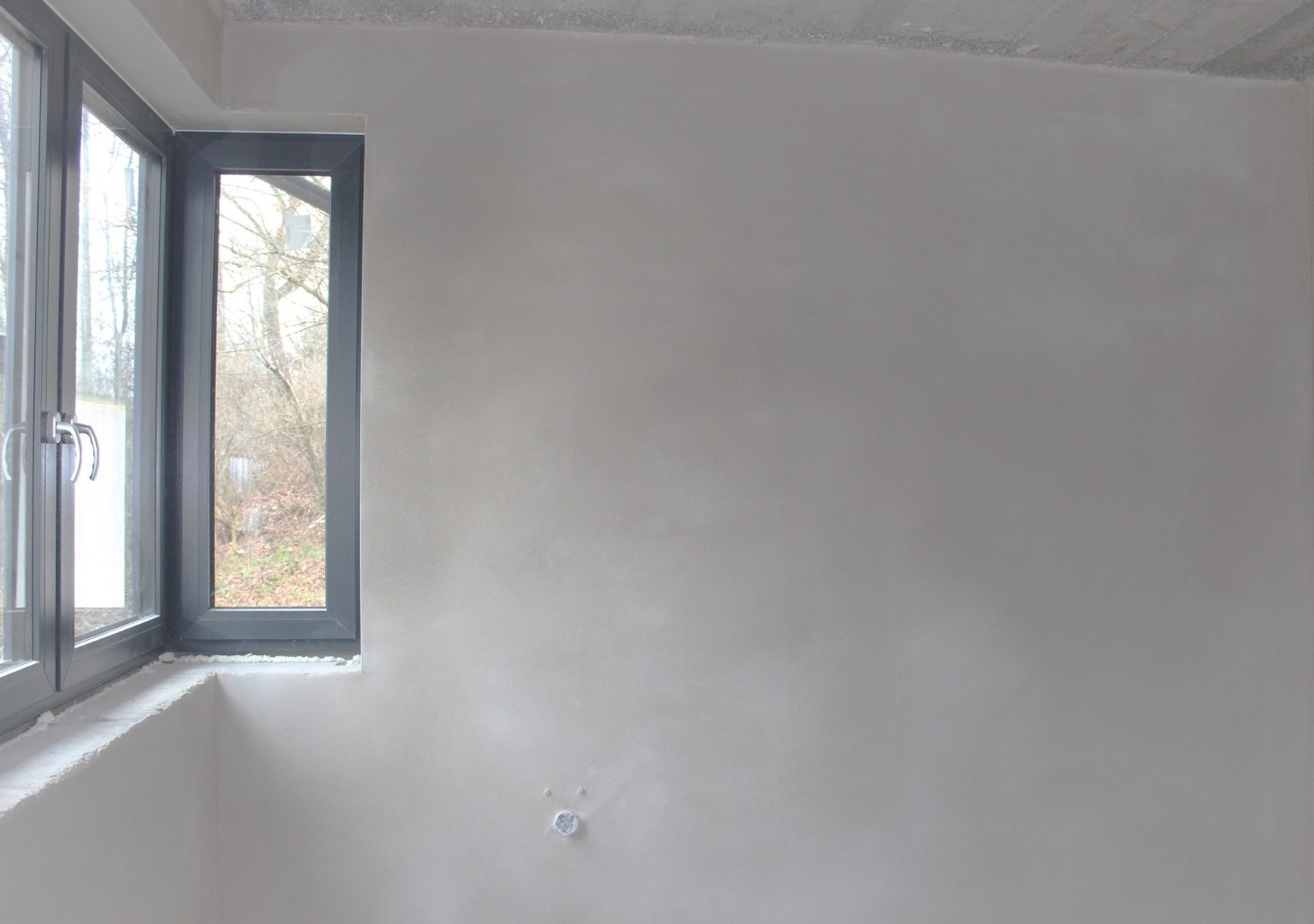 Świeżo nałożona, jasnoszara gładź na ścianie w pokoju, widoczne szare okno z widokiem na drzewa, surowy beton na suficie i wystający element instalacji elektrycznej przy podłodze.