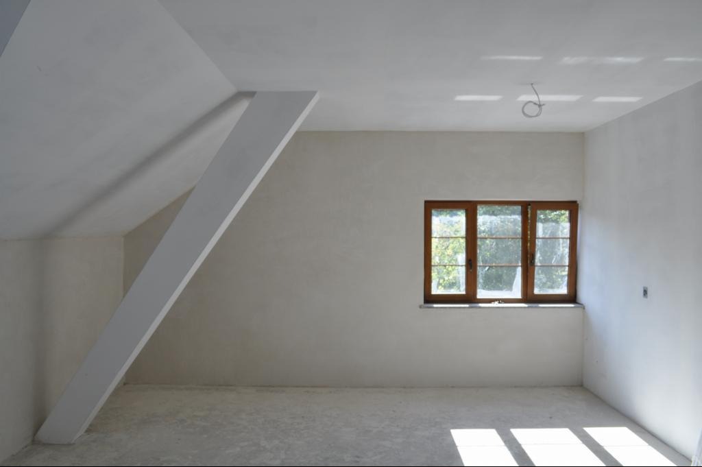 Poddasze po tynkowaniu maszynowym, widoczne świeże, jasne tynki na ścianach i suficie, drewniane okno z widokiem na zieleń, oraz ukośny element konstrukcyjny.