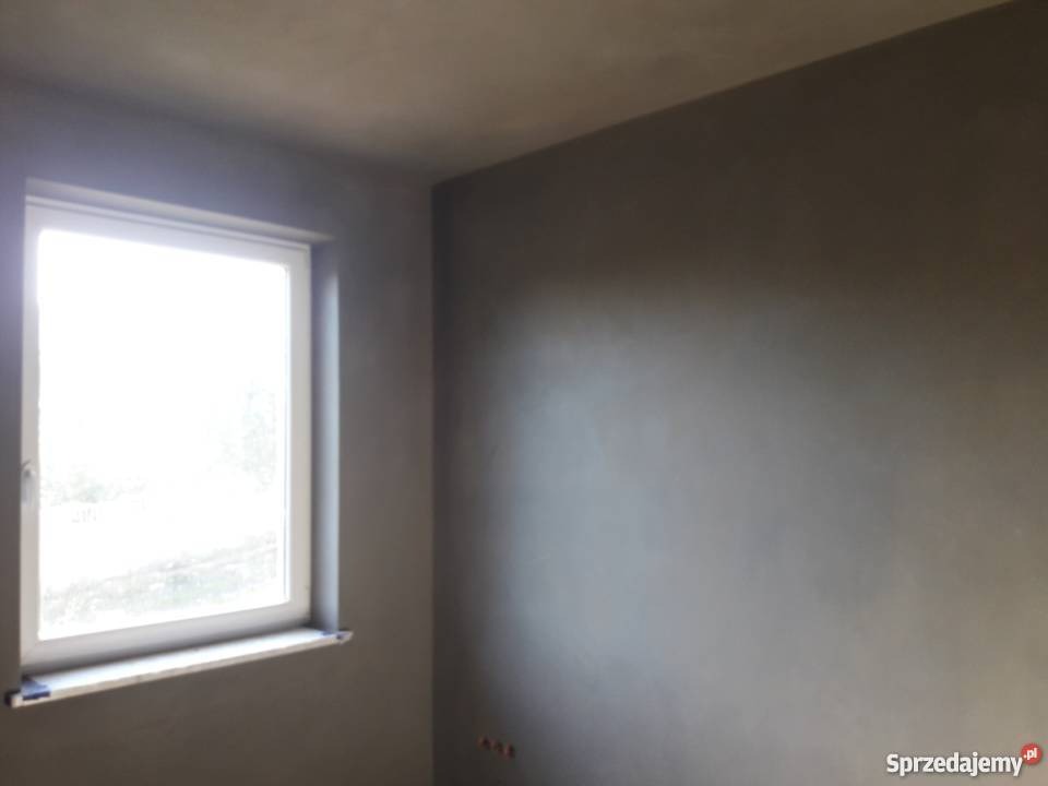 Świeżo nałożony, szary tynk na ścianach w pokoju z oknem, widoczne niedoskonałości faktury przed dalszą obróbką.
