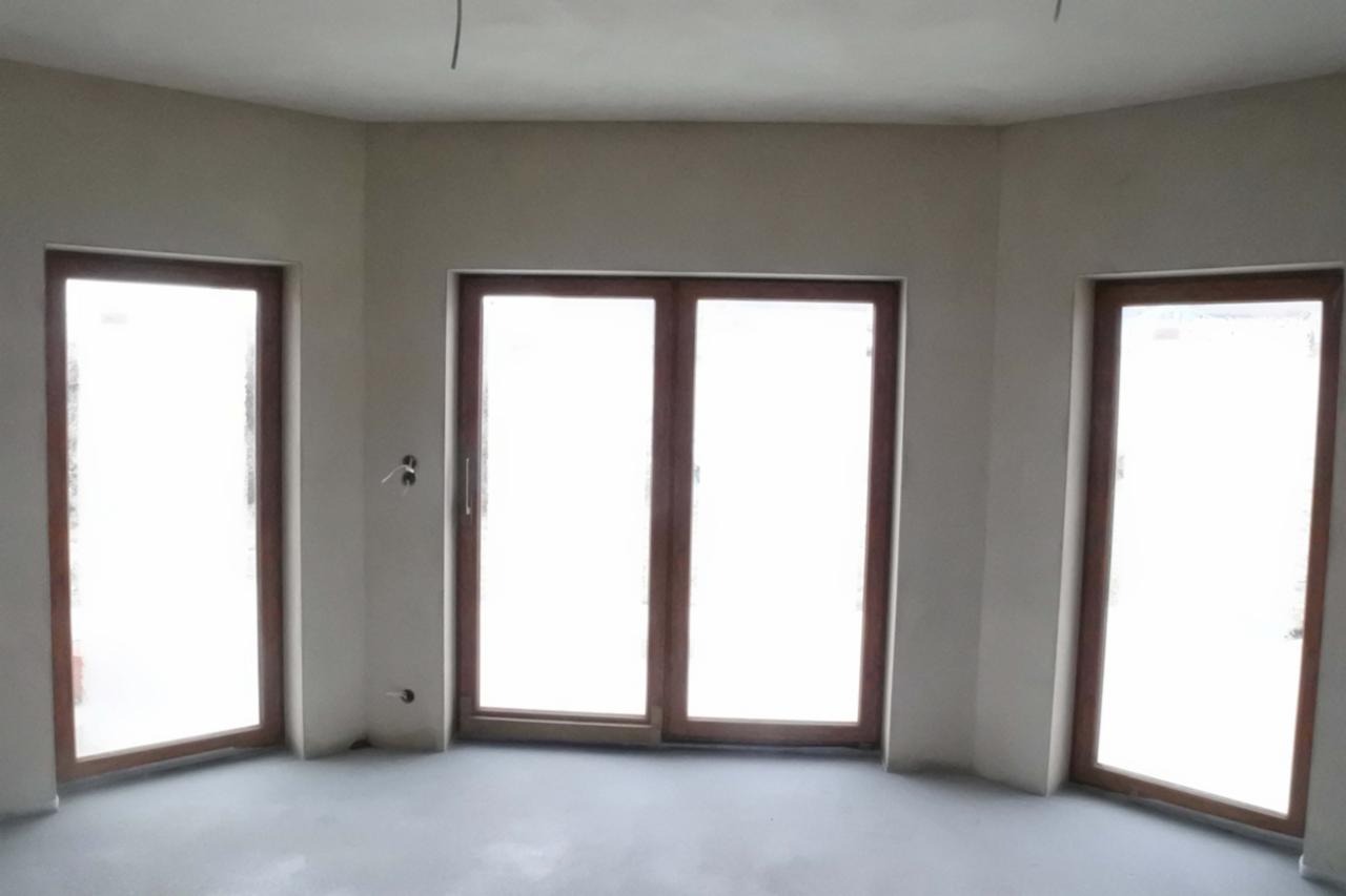 Pokój z trzema oknami w drewnianych ramach, ściany po nałożeniu gładzi, widoczne wystające kable i puszki elektryczne.