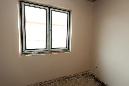 Pokój w trakcie prac wykończeniowych, ściany świeżo pokryte gładzią, okno z brązową ramą, surowa podłoga przed położeniem posadzki, widoczne ślady po instalacjach elektrycznych.