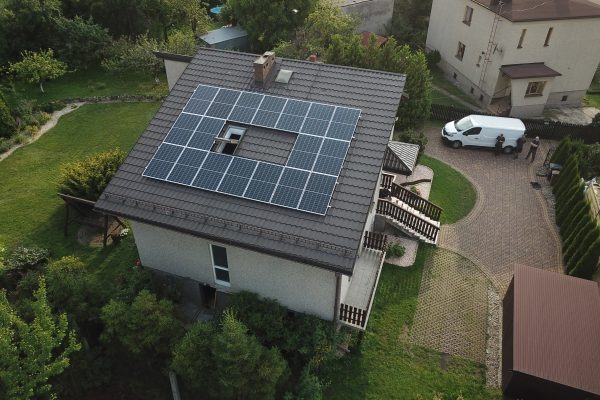 Widok z góry na dom z panelami słonecznymi na dachu, pozostawiającymi wolną przestrzeń wokół okna dachowego, z białym vanem i dwoma osobami w tle.