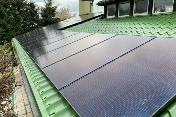 Panele słoneczne na zielonym, metalowym dachu domu z kominem, widok z poziomu gruntu.