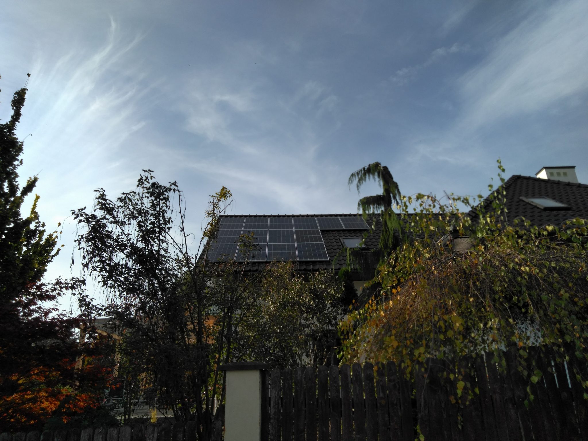 Panele fotowoltaiczne na dachu domu jednorodzinnego, widoczne zza drewnianego płotu i drzew w jesiennej scenerii, z delikatnymi chmurami na niebie.