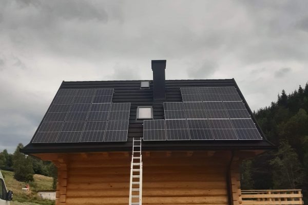 Panele słoneczne na dachu drewnianego domu z kominem i oknem dachowym, drabina oparta o dach, pochmurne niebo w tle.