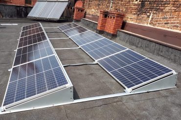 Dwa rzędy paneli słonecznych zamontowane na płaskim, bitumicznym dachu, z widocznym kominem z czerwonej cegły i kolektorem słonecznym w tle.