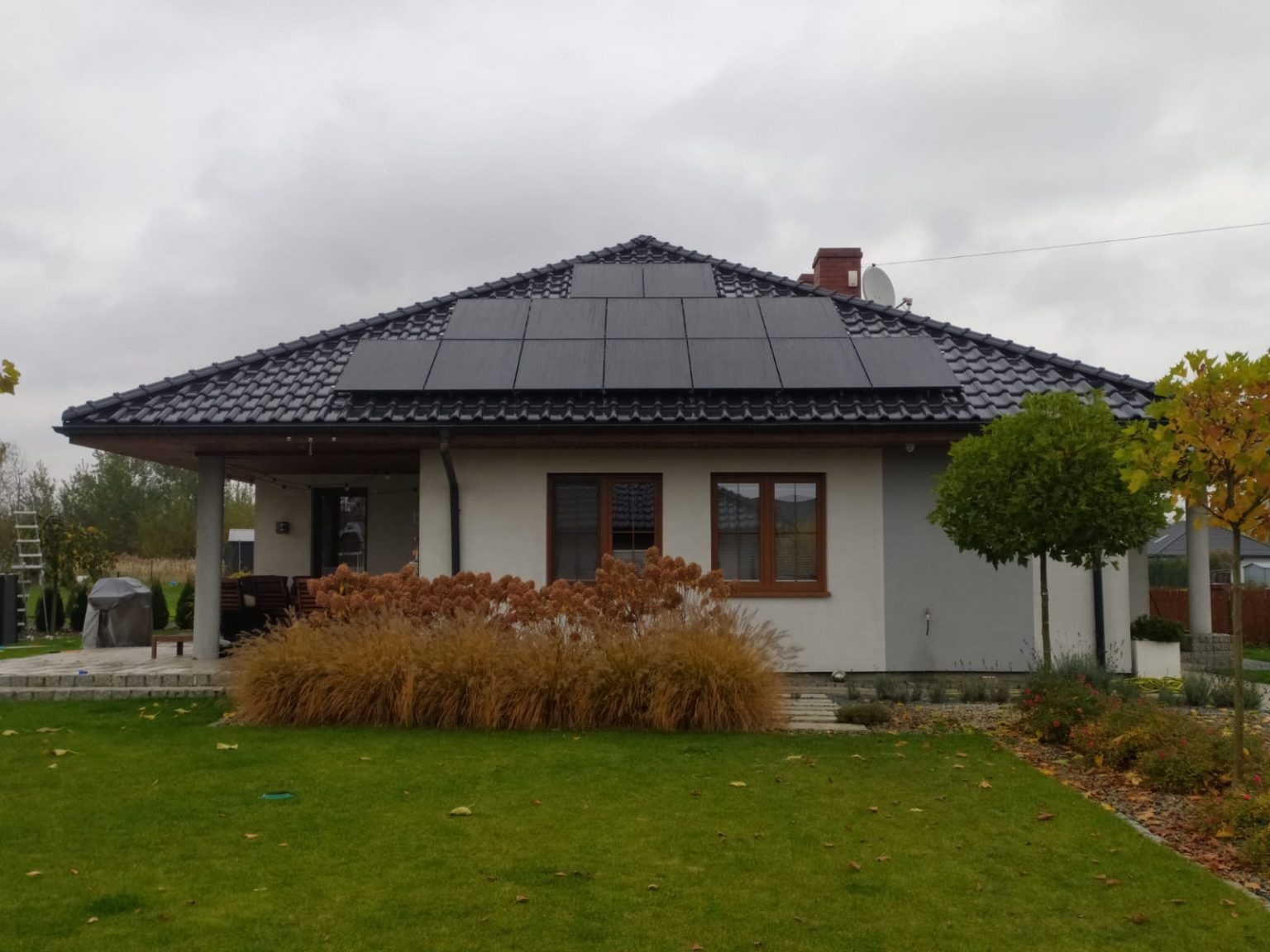 Dom jednorodzinny z panelami słonecznymi na dachu pokrytym czarną dachówką, widok z ogrodu w pochmurny dzień.