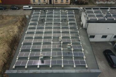 Widok z góry na instalację paneli słonecznych na płaskim dachu budynku przemysłowego, widoczne elementy konstrukcyjne i okablowanie, zarys drzew i budynków w tle.