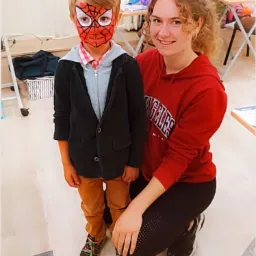 Chłopiec z pomalowaną twarzą w stylu Spider-Mana, ubrany w marynarkę, pozuje z uśmiechniętą kobietą w czerwonej bluzie z kapturem w jasnym pomieszczeniu.