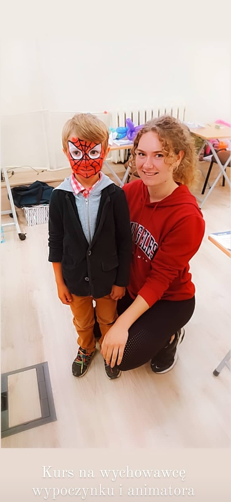 Chłopiec z pomalowaną twarzą w stylu Spider-Mana, ubrany w marynarkę, pozuje z uśmiechniętą kobietą w czerwonej bluzie z kapturem w jasnym pomieszczeniu.