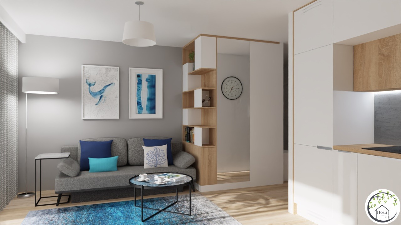 Nowoczesny salon z szarą sofą, niebieskimi poduszkami, okrągłym stolikiem kawowym na tle ściany z akwarelami i regałem z jasnego drewna, z widokiem na białą zabudowę kuchenną.