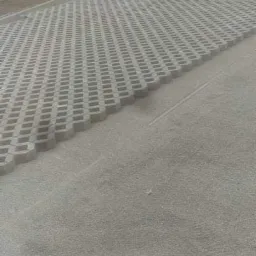 Ułożona ażurowa kostka betonowa na podsypce z kruszywa, widoczne obrzeże, w tle trawnik i drzewa.