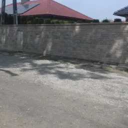 Długi, szary, betonowy mur z regularnych bloków, z metalowymi drzwiczkami rewizyjnymi po lewej stronie, oddzielający posesję z domem o czerwonym dachu od drogi gruntowej.