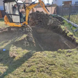 Żółta mini koparka JCB 8015 wykopuje głęboki dół w ziemi na trawiastym terenie, z widoczną warstwą gliny i gruzu, w tle ogrodzenie panelowe i budynki mieszkalne